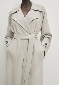Beżowy płaszcz trench z szerokim kołnierzem, długimi rękawami i paskiem. Posiada dwie przednie kieszenie oraz guziki przy mankietach; gładka tekstura.
