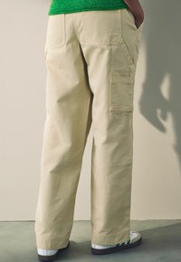 Beige katoenen cargobroek met zijzakken, losse fit, opvallende stikdetails en een opgerolde zoom. Draagt met witte sneakers.