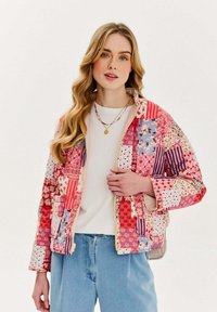 Prešívaná bunda s patchwork dizajnom v červených a ružových odtieňoch, s kvetinovými a pruhovanými vzormi, so zipsom vpredu a širokými rukávmi.