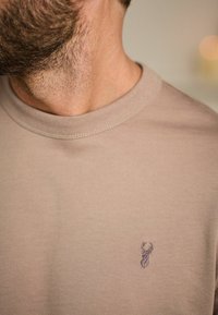 T-shirt à manches courtes de couleur tan, fabriqué en tissu doux, avec un logo de chevreuil subtil brodé sur la poitrine gauche et un col côtelé.