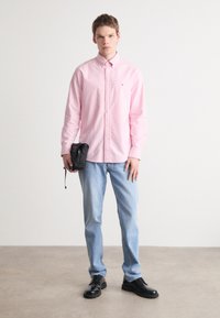 Gaiši rozā krekls ar pogām, gaiši zilas džinsu bikses, melni apavi, tur rokā mazu melnu somu. Redzams gluds audums un pielāgotas siluets.