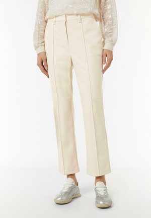 Cream-colored tailored pants with front pleats, flat front, and a straight cut. Paired with silver sneakers, showcasing a casual look.