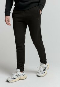 Zwarte, taps toelopende sweatpants van een gladde stof, met zijzakken en een subtiele hexagonale logo. Gestyled met witte en zwarte sneakers.