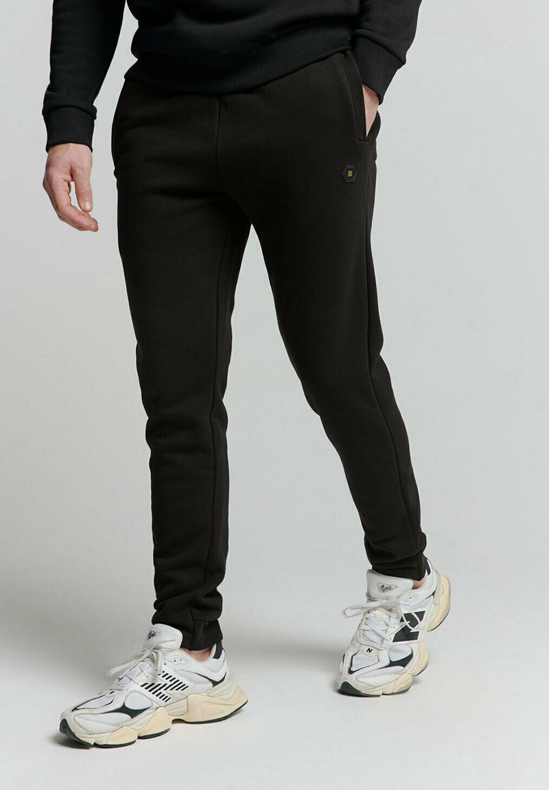 Zwarte, taps toelopende sweatpants van een gladde stof, met zijzakken en een subtiele hexagonale logo. Gestyled met witte en zwarte sneakers.