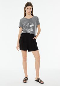 T-shirt grigio in cotone con grafica "Cartolina dal Paradiso", abbinato a pantaloni corti plissettati neri e sandali neri con cinturini. Sfondo bianco.