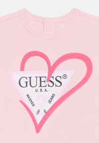 Camiseta de algodón rosa con un gran gráfico de corazón en rosa alrededor de un logotipo en forma de triángulo con "GUESS U.S.A." y el texto: "JEANS LAVADOS."