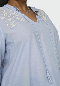 Világoskék blúz virágos hímzéssel, V-kivágással, zsinóros megoldással bojtokkal, puha anyagból és laza fazonban.