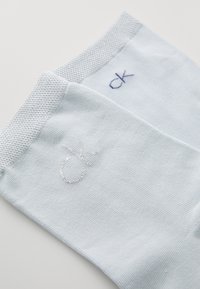 Gaiši zili zeķes ar adītām aprocēm, uz mīkstas ribota auduma redzami sudraba un tumši zili "ck" logotipi.