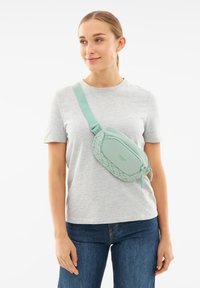 Borsa a tracolla verde menta con un design geometrico testurizzato, tracolla regolabile e scomparti con zip, indossata sopra una maglietta grigia.