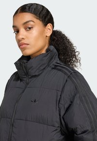 Γυναίκα με σγουρά μαλλιά μαζεμένα, φορώντας μια μαύρη пухέρ jacket με ψηλό γιακά και λογότυπο Adidas στο στήθος, καθώς και ρίγες στα μανίκια.