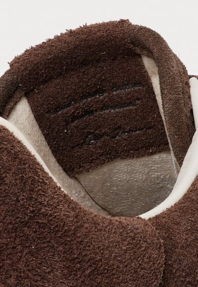 Pantofole in suede marrone con una texture morbida e pelosa, fodera bianca e una forma arrotondata con risvolto. Dettagli di cucitura sottili sono visibili.