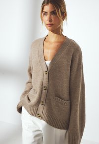 Beige oversized cardigan i mjukt stickat material, med V-ringning, fem knappar, två framfickor samt ribbade mudd- och nederkanter.