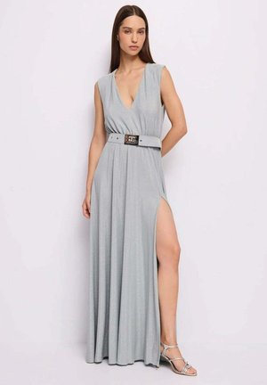 Donna in piedi che indossa un abito lungo argento senza maniche con scollatura profonda a V, vita con cintura, spacco alto sulla coscia e sandali argento con cinturini su sfondo semplice.