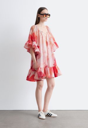 Žena v nadrozmerných kvetovaných šatách s volánikovými rukávmi a lemom, veľké slnečné okuliare a biele tenisky s čiernymi pruhmi, stojaca z boku.