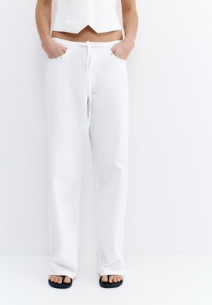 Nedre torso iført hvide buksesnøre med lommer, hænder i lommerne og sorte åbne tåsandaler på en hvid baggrund.