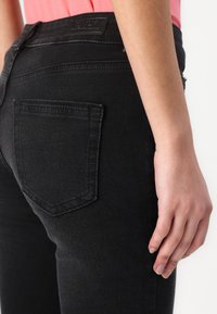 Black denim jeans featuring a back pocket with stitching details and a branded label at the waistband, shown from the side.