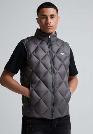 Giovane uomo che indossa un gilet imbottito grigio sopra una maglietta nera a maniche corte, in piedi con le mani nelle tasche del gilet.