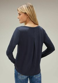 Marineblaues Langarmshirt mit Rundhalsausschnitt und Rückenfaltendetail, kombiniert mit blauen Jeans. Glattes Material und taillierte Form.