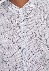 Biała koszula z guzikami, z czarnym geometrycznym wzorem z drobnymi liniami i kropkami, podkreślająca fakturowany materiał i kołnierzyk.