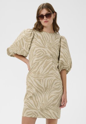 Frau trägt ein beiges und cremefarbenes Zebra-Muster Puffärmelkleid und dunkle, übergroße Sonnenbrille, posiert mit der Hand in der Hüfte vor schlichtem Hintergrund.