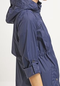 Imperméable bleu marine avec fermeture éclair, capuche ajustable, taille élastique et poignets boutonnés. Tissu lisse et résistant à l'eau.