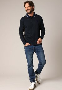 Polo à manches longues bleu marine avec un col contrastant, assorti à un jean bleu et des baskets blanches, présenté sur un modèle masculin debout.