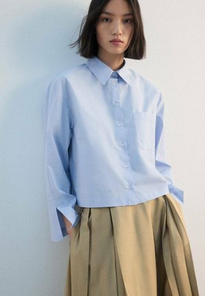 Camisa azul claro de botones con cuello, mangas largas y un bolsillo en el pecho; combinada con una falda plisada beige.