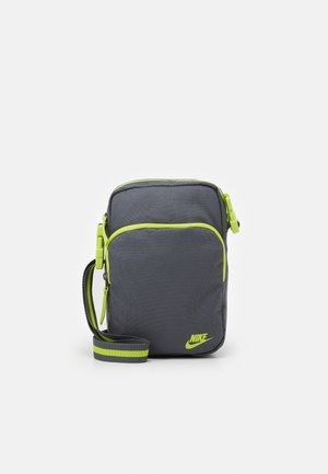 Bolso bandolera pequeño de Nike en gris oscuro con cremallera de borde verde brillante, logo y correa ajustable a rayas, colocado de pie sobre un fondo liso.