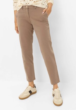 Person trägt hellbraune, gerade Hosen, beige Sneakers mit schwarzen Streifen und ein leichtes Strickoberteil mit Cut-out-Muster.