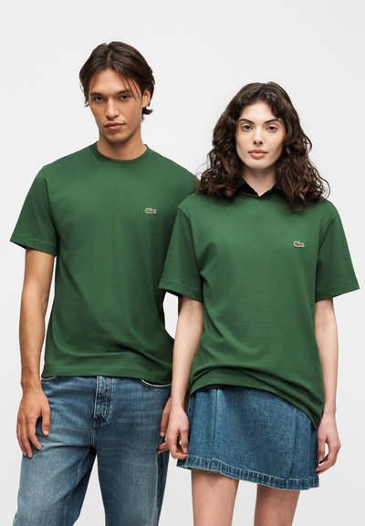 Мъж и жена с еднакви зелени тениски Lacoste; мъж в сини дънки, жена в синя дънкова пола, застанали пред едноцветен фон.