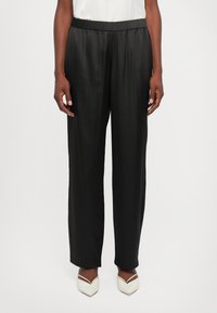 Persona, kas valkā melnu elastīgu jostasvietas taisnā stila bikses un baltus ar smailiem purngaliem apavus, stāv uz vienkārša balta fona.