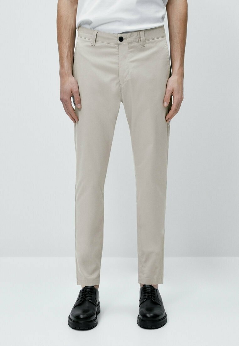 Homme portant un pantalon slim beige avec un bouton noir et des chaussures noires à lacets, debout devant un fond blanc uni.