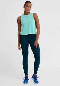 Hellgrünes ärmelloses Croptop, kombiniert mit dunkelgrünen Leggings und blauen Sportschuhen. Glatter Stoff, figurbetontes Design und minimalistischer Stil.