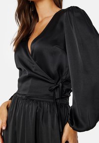 Femme portant un chemisier noir portefeuille avec de longues manches bouffantes et une taille nouée, assorti d'une jupe noire coordonnée.