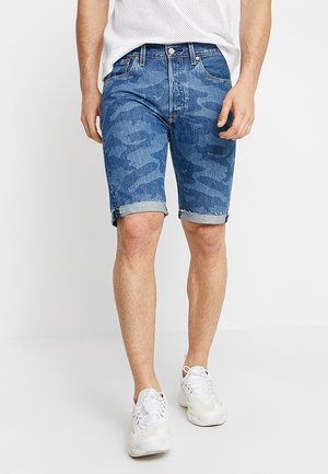 Homme portant un short en jean à motif camouflage bleu avec ourlets retroussés, un haut blanc sans manches en mesh, et des baskets blanches sur fond blanc uni.