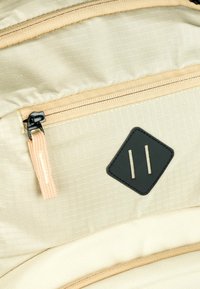 Sezione di uno zaino in tessuto beige con una tasca diagonale con zip, materiale testurizzato e una patch in pelle nera con due linee verticali.
