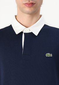 Jūras spēku zils polo krekls ar baltu apkakli, divu pogu pogāju un mazu zaļu krokodilu logotipu kreisajā krūtī. Izgatavots no kokvilnas materiāla.