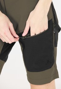 Olivgröna shorts har en svart dragkedjeficka och kontrasterande tygpaneler. Materialet är lätt och har en jämn textur.