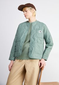 Gequilted groene jas met drukknopen, een voorzak en textiel met patroon, gedragen over een mintgroene trui en beige broeken. Bruine pet op het hoofd.
