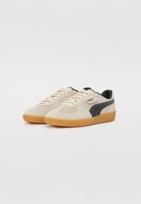 Beżowe zamszowe sneakersy z czarnymi akcentami, okrągłym noskiem i gumową podeszwą. Posiadają sznurowadła i metkę na języku.