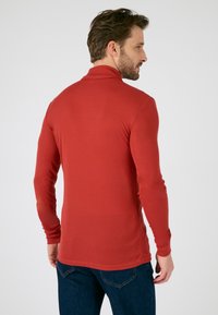 Red long-sleeve polo shirt with a mock neck, slim fit, ribbed texture, and a rounded hem, paired with dark blue jeans.