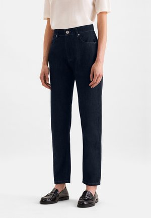 Persoon draagt donkerblauwe jeans met rechte pijpen, een wit T-shirt met korte mouwen en zwarte leren loafers, staand tegen een witte achtergrond.