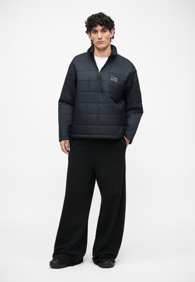 Black, quilted pullover with a half-zip and front pocket; worn over white shirt. Loose-fitting black pants and black shoes complete the look.