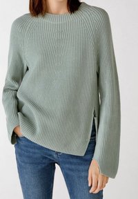 Femme portant un pull en maille côtelée vert clair avec des manches larges et un détail de fermeture éclair sur le côté, associé à un jean bleu, main dans la poche.