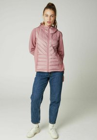 Lyserød quiltet jakke med hætte, der har lodrette ribbede ærmer og lynlås foran. Båret over blå jeans og hvide sneakers.