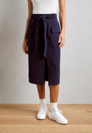 Femme portant une jupe bleu marine longueur genou avec des poches frontales et une ceinture nouée à la taille, associée à des baskets blanches et des chaussettes sur un sol en bois.