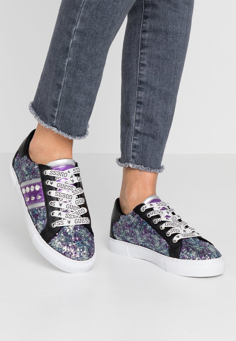 Personne portant des baskets multicolores étincelantes avec des lacets blancs de marque "GUESS" et un jean gris foncé aux ourlets effilochés.