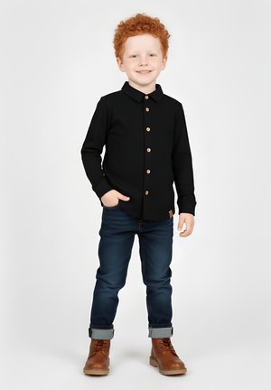 Czarna koszula z długim rękawem i guzikami, noszona z ciemnoniebieskimi dżinsami i brązowymi butami. Strój posiada kołnierz i detal z naszywką z tkaniny.