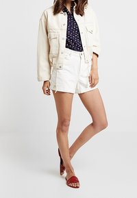 Ljusblå denimjacka, marinblå skjorta med små mönster, vita shorts och röda sandaler med remmar. Avslappnad passform med synliga sömmar.