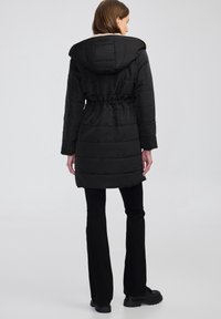 Černé, vycpané vnější oblečení s kapucí, staženým pasem a strukturovanou tkaninou. Má dlouhé rukávy a sahá do poloviny stehna. Ležérní design.